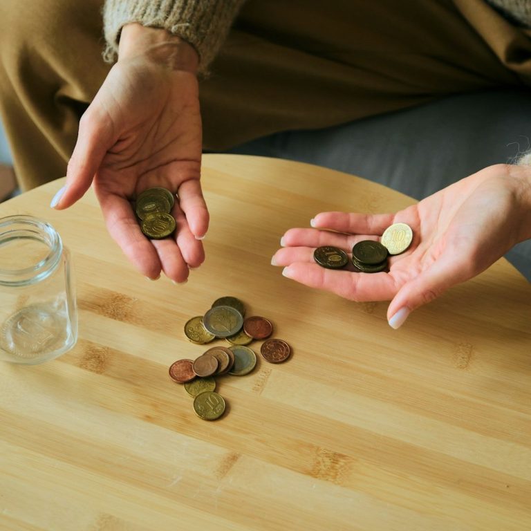 Costes Manos sosteniendo monedas, con algunas monedas dispersas sobre una mesa y un frasco vacío.